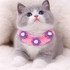 A grey and white kitten wearing a pink crochet bib decorated with purple and white flowers, set against a soft, light background.