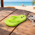 A bright green thongs shape keyring rests on a wooden table, with keys nearby and a beach scene in the background.