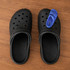 A pair of black clog shoes with a single blue charm positioned on one of the shoes.