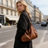 A brown polyurethane tote bag is worn over the shoulder by a woman in a city street setting.