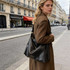 A black polyurethane tote bag is being carried by a model dressed in a brown coat on a Parisian street.