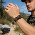 A paracord bracelet in brown with a black clasp, worn by a man outdoors on a hiking trail. A close-up of the clasp is included.