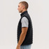 A man in a side profile wearing a black puffer vest over a grey t-shirt. The vest has a logo on it.
