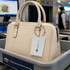 A cream handbag sits in a security tray with a luggage tag attached, featuring a logo and silver hardware.