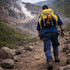 A hiker wearing a yellow backpack walks along a rocky trail surrounded by greenery and mountains in the background.