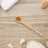A natural wooden face brush with a beige bristle head, set against a wooden background with soft towels and seashells.