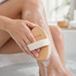 A wooden brush with a light-coloured handle is used on a leg with soap lather in a bath setting.