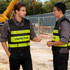 Two men wearing black and hi-vis yellow reflective safety vests with a logo visible, standing on a construction site.