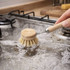 A wooden cleaning scrubber with a long handle and natural bristles, submerged in bubbly water on a stove.