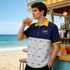 A man wearing a blue and yellow Hawaiian shirt with a patterned design stands by a beachside bar, holding a drink.
