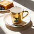 A gold ceramic coffee mug with a colourful bird design, paired with a matching saucer, sits beside toast and blueberries.