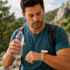 A man in a blue shirt holds a water bottle while looking at a green and pink Medicine Box Wristband on his wrist.