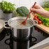 A wooden spoon with a long handle in a pot of boiling water, surrounded by fresh vegetables on a kitchen countertop.