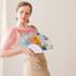 A paper hand-held fan featuring an abstract colourful design, held by a woman wearing a pink blouse and beige skirt.