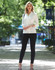 A mid sleeve knit top in white, paired with black leggings, set against a green park backdrop. The model carries a bag.
