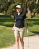A woman walks on a golf course wearing a black polo shirt and beige shorts, holding a golf club.