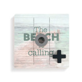 A square wood board for tic tac toe with a lake pier scene that says "The Beach is Calling" with a gray O and black X on top.