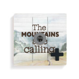 A square wood board for tic tac toe with a mountain scene that says "The Mountains are Calling", with a gray O and black X on top.