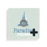 A light green square board with a sailboat and the saying "Paradise Found" for tic tac toe with a gray O and black X on top.