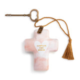 A light pink cross shaped sculpture with a heart in the center that says "comfort and peace" with a gold tassel and metal key attached.