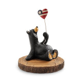 A black bear sitting on a wood stump and holding a heart shaped balloon of the American flag.