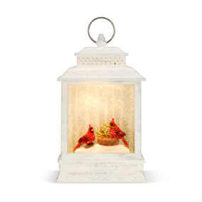 An illuminated white washed wooden musical lantern, with two red cardinals, a nest, and faux snow enclosed. With a silver ring.