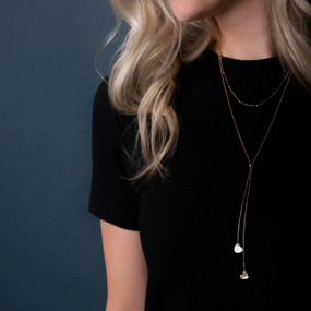 Close up of a woman wearing a black top and a long gold chain lariat necklace with two heart charms at the bottom.