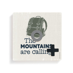 A light wood square board with a green backpack and the saying "The Mountains are calling" for tic tac toe with a gray O and black X on it.