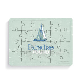 A rectangular 24 piece wood postcard puzzle with a sailboat on a light green background with the saying "Paradise Found".