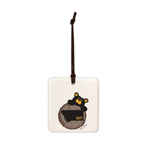 A white square hanging ornament with a black bear peeking over a tree stump with Montana on it.