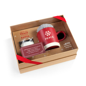 A brown gift box wrapped with a red ribbon that includes a red and white mug that says "Peace", a pair of cozy knit socks and a soy blend candle in a glass holder.