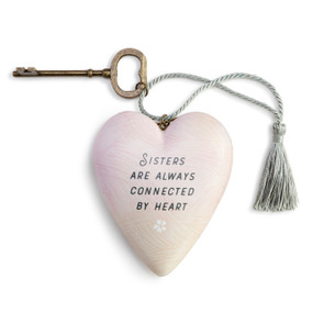 Heart shaped sculpture with a silver tassel and metal key attached. The heart is light pink with a white flower and says "Sisters are Always Connected by Heart".