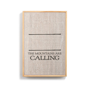 A light wood framed wall art that says "The Mountains are Calling" on a wood grain background under two black lines for personalization.