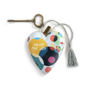 A heart shaped sculpture with a gold key and silver tassel featuring artwork created by ArtLifting artist Susan Spangenberg of different colors and sizes of circles overlapping and says "Enjoy the Joy".