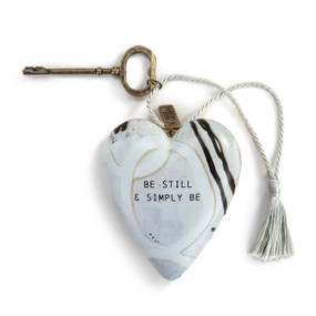 A heart shaped sculpture with a gold key and silver tassel featuring artwork created by ArtLifting artist Cheryl Kinderknecht. The image is a gray background with black lines and circles and says "Be Still & Simply Be".
