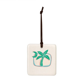 A square hanging tile ornament with a graphic image of a green moon and palm tree on a sand colored background.