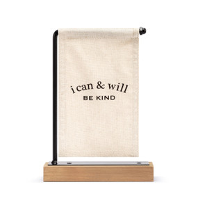 A cream canvas sign with black writing that says "i can & will Be Kind" hanging on a black metal stand with a wood base.