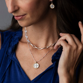 A woman in a blue blouse wearing a layered silver necklace and earrings each with a round silver cross medallion.
