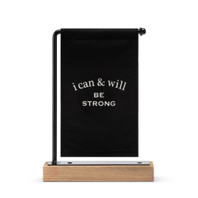 A black canvas sign with white writing that says "i can & will Be Strong" hanging on a black metal stand with a wood base.