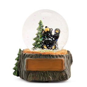 A glass snow globe with a sitting black bear next to a pine tree. The base is sculpted like a tree trunk and has a rectangular space for personalization.