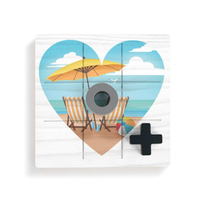A square white wood board for tic tac toe with heart shaped graphic artwork of two beach chairs and an umbrella at the beach, displayed with a gray O and black X on top.