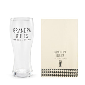 A clear pilsner glass that says "Grandpa Rules The Grill 'N Chill" next to a cream towel with the same saying and a houndstooth pattern at the bottom.