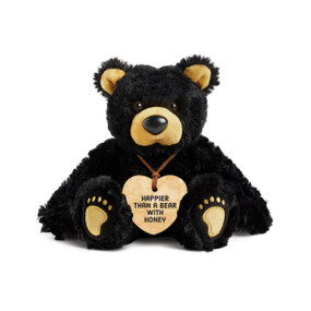 A black plush bear with brown ears, nose and paws. The bear is wearing a heart shaped yellow painted wood ornament around its neck that says "Happier Than A Bear With Honey".