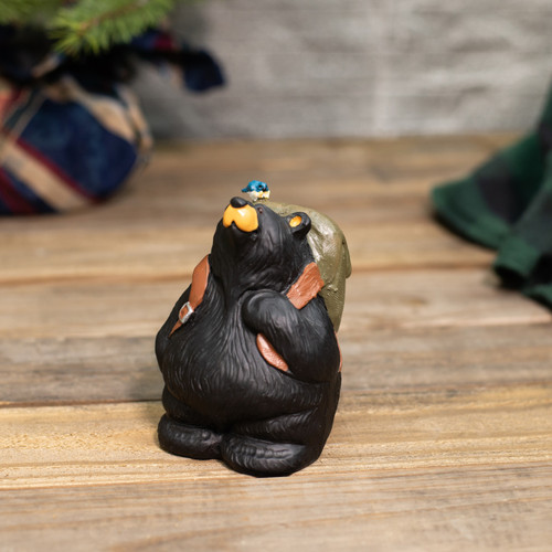 A carved figurine of a black bear wearing a backpack with a little bluebird perched on top, displayed on a wood table.