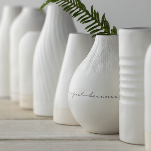 A close up image of several white vases with various shapes, sizes, and textures. One vase is filled with a stem of greenery.