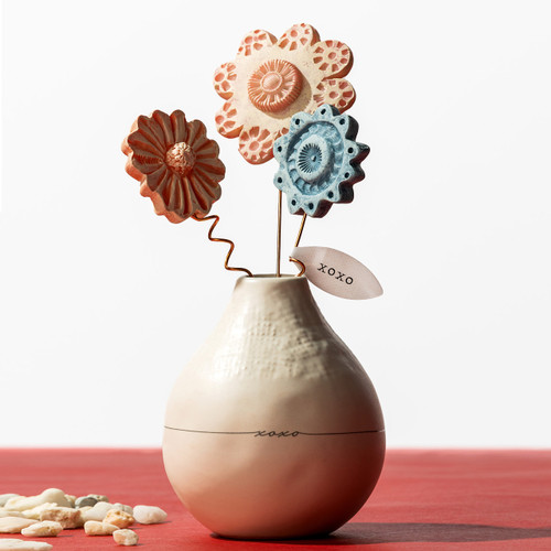 A cream stoneware vase with a black line around the middle that says "xoxo". The vase is holding three pink and blue decorative resin flowers on copper stems.