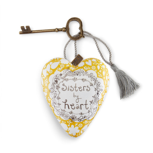 A white and yellow heart shaped sculpture that says "Sisters by heart" with a silver tassel and metal key attached.