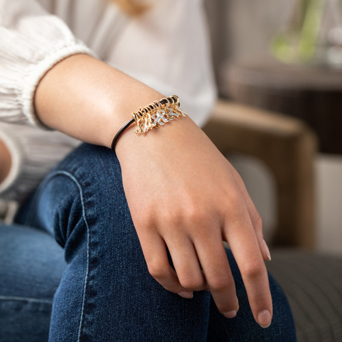 A woman in a white blouse and blue jeans displaying a black cord bracelet with several gold and silver charms.