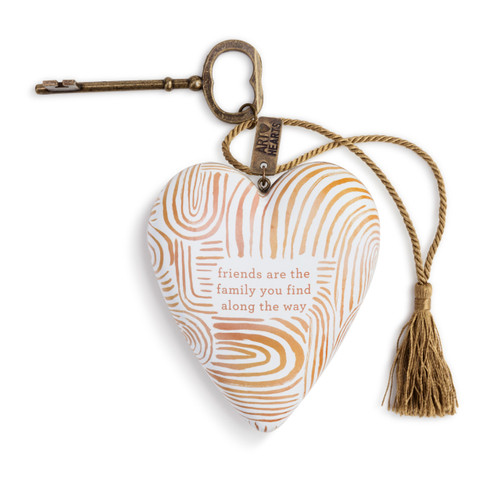 A white art heart with abstract orange stripes, and reads friends are the family you find along the way" in the center. With a bronze key and a gold tassel."