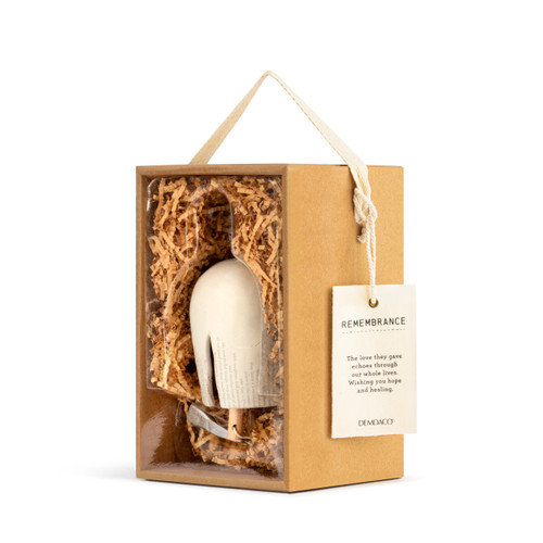 A left facing view of a cream Inspired Bell" with a single crack, small gray font coming from the bottom edge, a twine rope, and silver and wooden beads. Placed in a brown cardboard box with brown crinkle paper, and an ivory tag that reads "Remembrance""